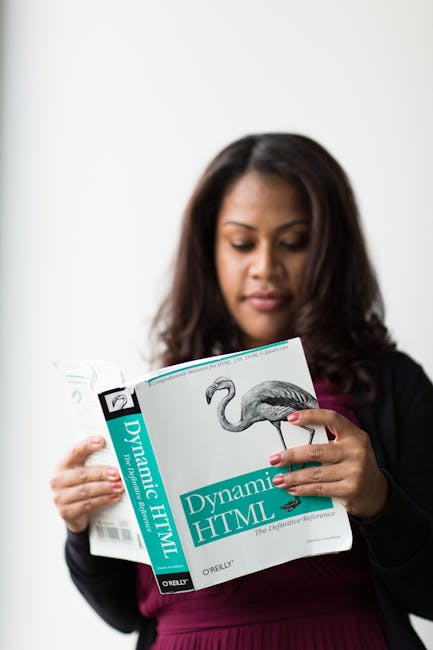 The image shows a woman with long, dark hair reading a gardening book titled 'Dynamic HTML' by O'Reilly. She is dressed in a dark blazer over a maroon top, standing indoors against a plain, neutral ba