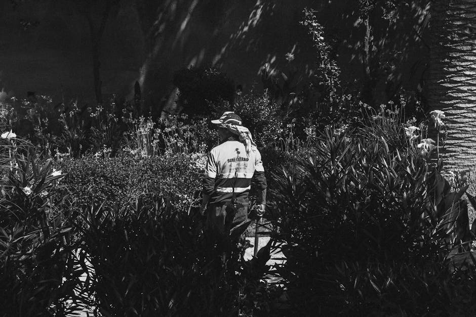 A gardener in a high-visibility jacket and wide-brimmed hat is working in a lush garden at night, surrounded by dense foliage, flowering plants, and shrubs. The scene reveals a well-maintained landsca